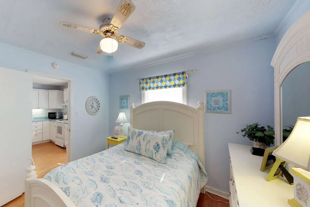 A cozy bedroom featuring a bed with decorative pillows, a ceiling fan, bedside lamps, colorful window treatments, and tasteful wall art. The room is bathed in gentle natural light, creating a peaceful atmosphere ideal for rest and relaxation.