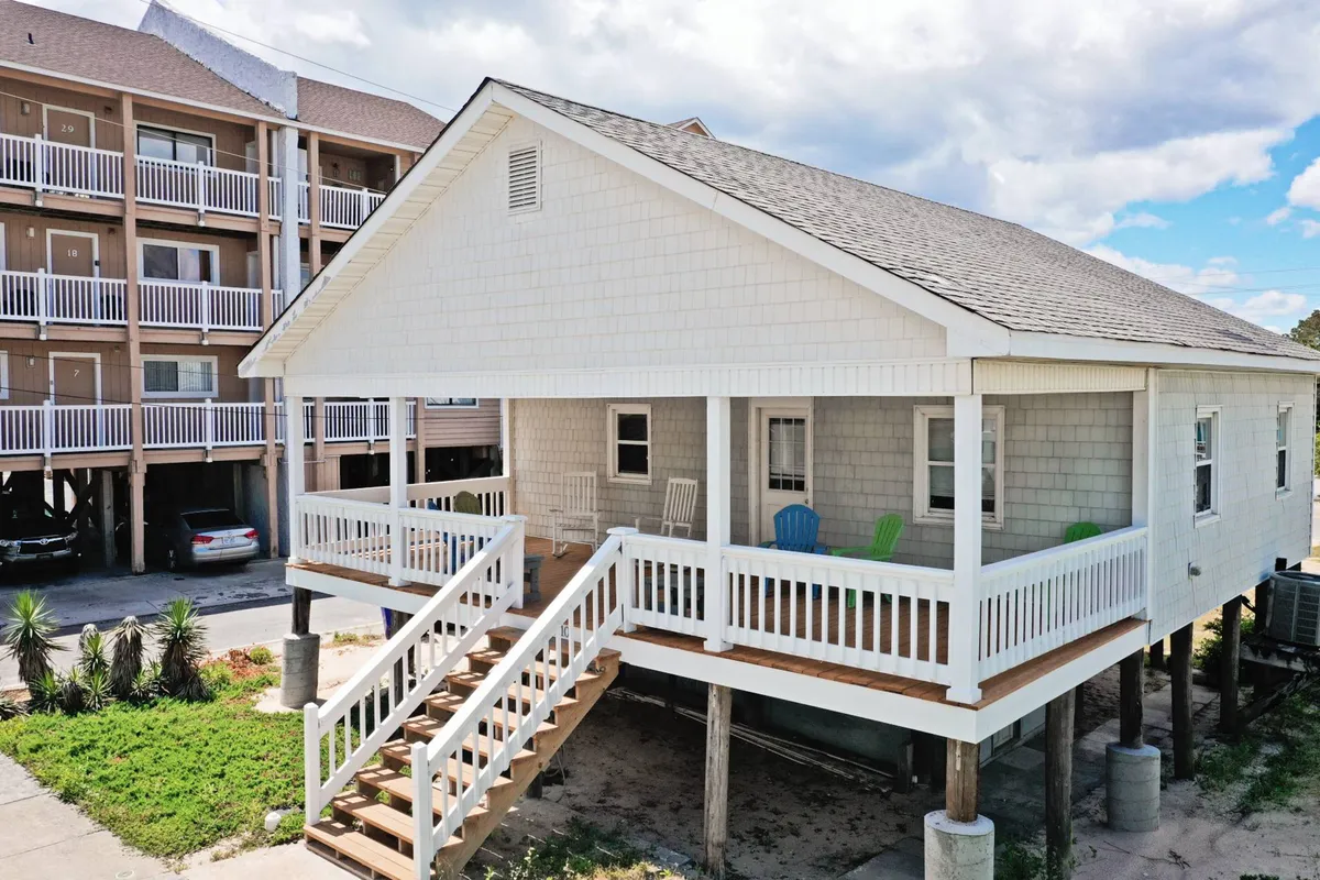 This exterior shot focuses on the large front porch of Hollis House, featuring colorful chairs and inviting wooden stairs. The elevated structure hints at ocean proximity, while the porch sets the scene for family gatherings, outdoor meals, and even a family pet lounging nearby.
