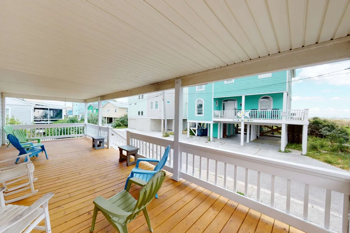 The image shows a charming wooden deck at Hollis House, furnished with inviting deck chairs that overlook Carolina Beach and the neighboring cottages. The golden glow of sunset bathes the space, suggesting opportunities for morning coffee or evening barbecues in a peaceful, oceanside setting.