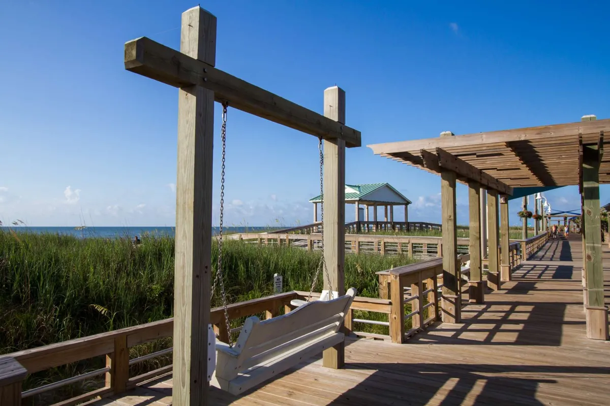This image features a serene exterior scene with a wooden swing overlooking a seaside gazebo, tall grass, and a glimpse of the ocean. The swing sits at the edge of a wooden walkway, inviting visitors to relax and enjoy the tranquil environment. The setting is intimate and peaceful, ideal for quiet moments together.