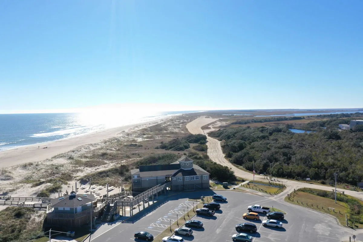 The image shows a relaxed exterior environment with the ocean, boardwalk, parking lot, and sand dunes. The boardwalk offers access to the beach while framing the ocean in the background, suggesting easy access to both vibrant beachfront activities and quiet moments together.
