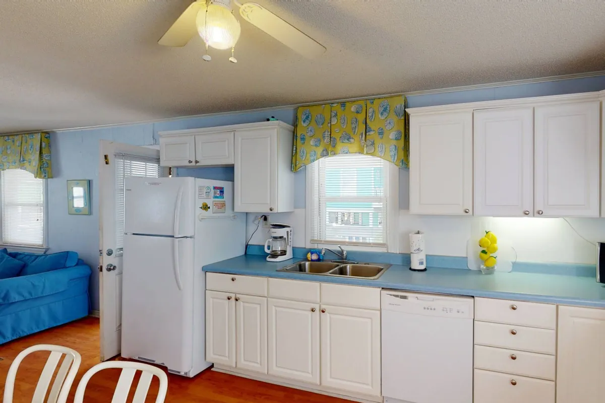 The image presents a welcoming kitchen and living space, featuring a blue couch, a kitchen counter, and a curtained window letting in soft daylight. The arrangement suggests comfort and the possibility of entertaining guests or spending quiet family time.
