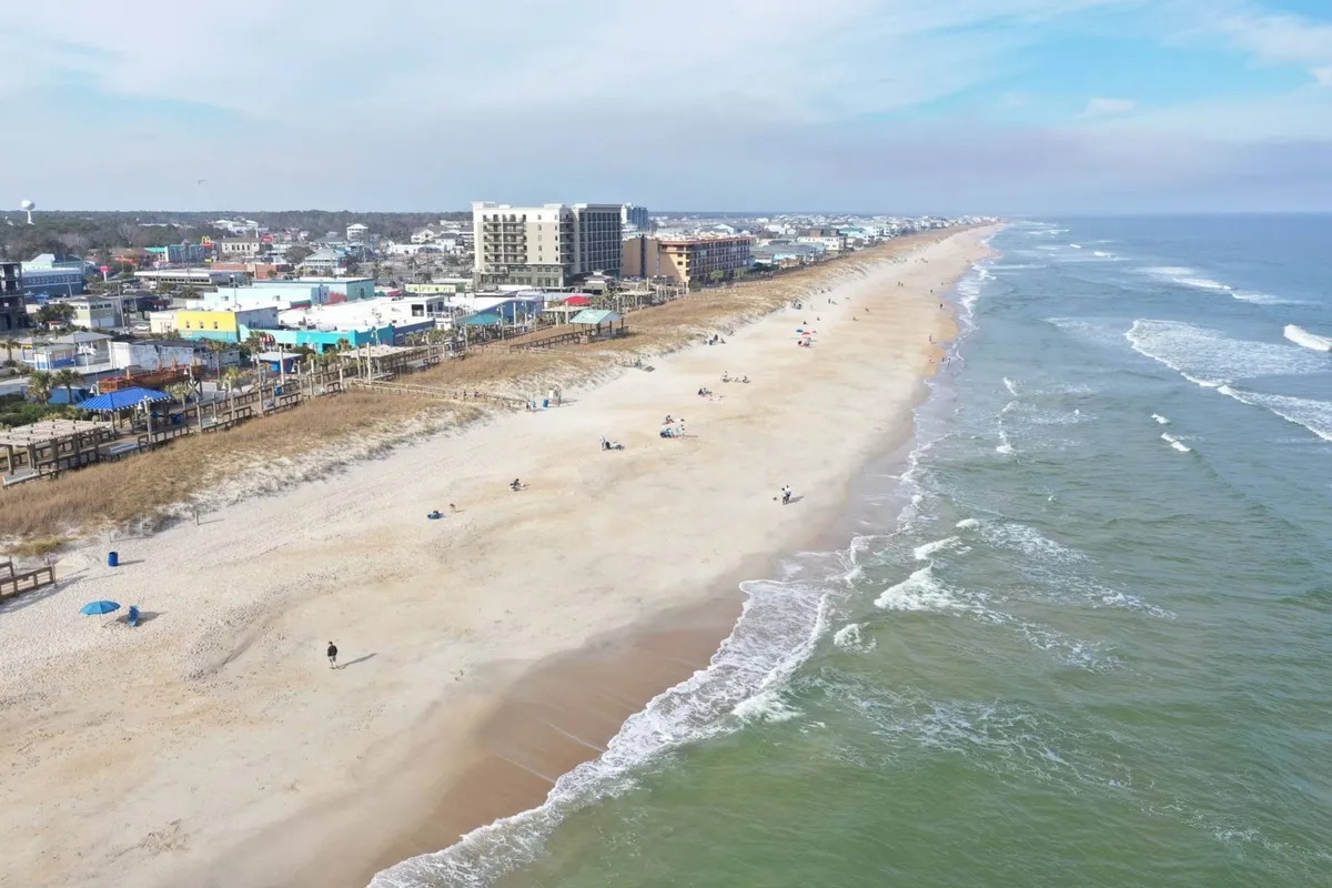 This exterior image captures a wide sandy beach stretching out to meet the gentle surf, with a boardwalk and nearby buildings in view. The scene includes children and families playing by the water, building sandcastles, and enjoying the beach—perfectly encapsulating the family-friendly, leisurely energy described in the section.