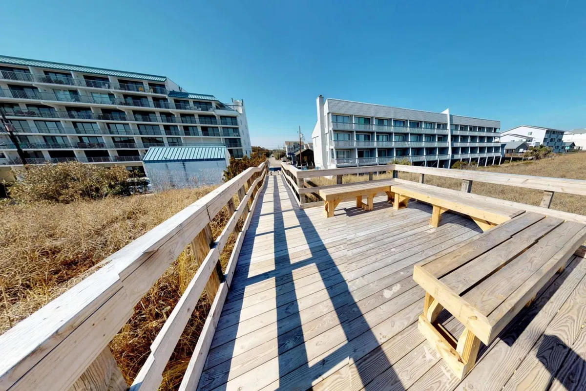 A welcoming wooden walkway lined by benches leads toward surrounding buildings, with sunset views and a lively, social atmosphere perfect for evening gatherings. The scene invites relaxation and group socializing, echoing the experience of starting the evening together before heading out.