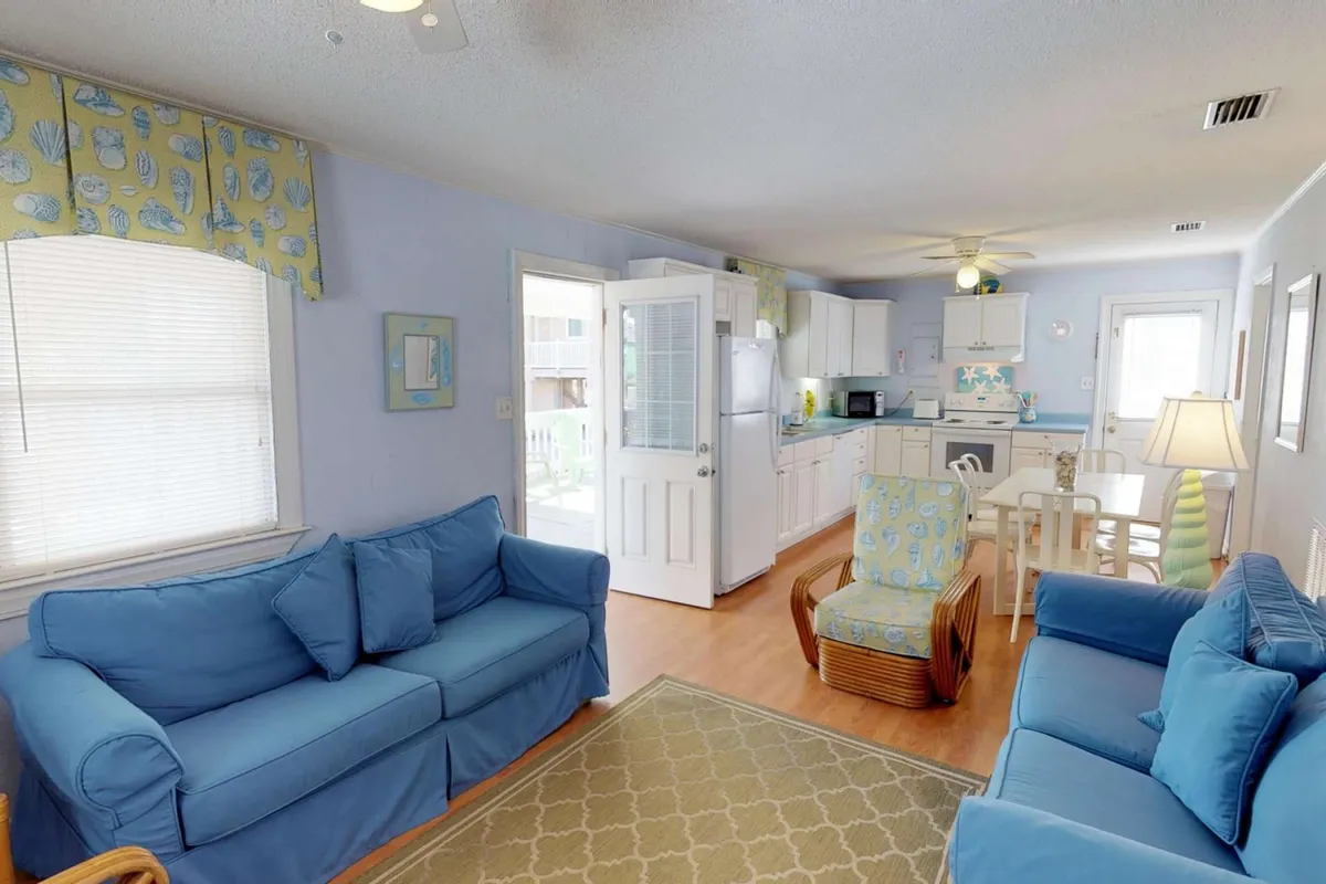 This photo captures the main living area with plush blue sofas, a central dining table, and an open kitchen in the background. A decorative lamp adds a homey touch and natural light streams in, enhancing the welcoming vibe. The layout clearly shows the seamless flow between the living and dining spaces.