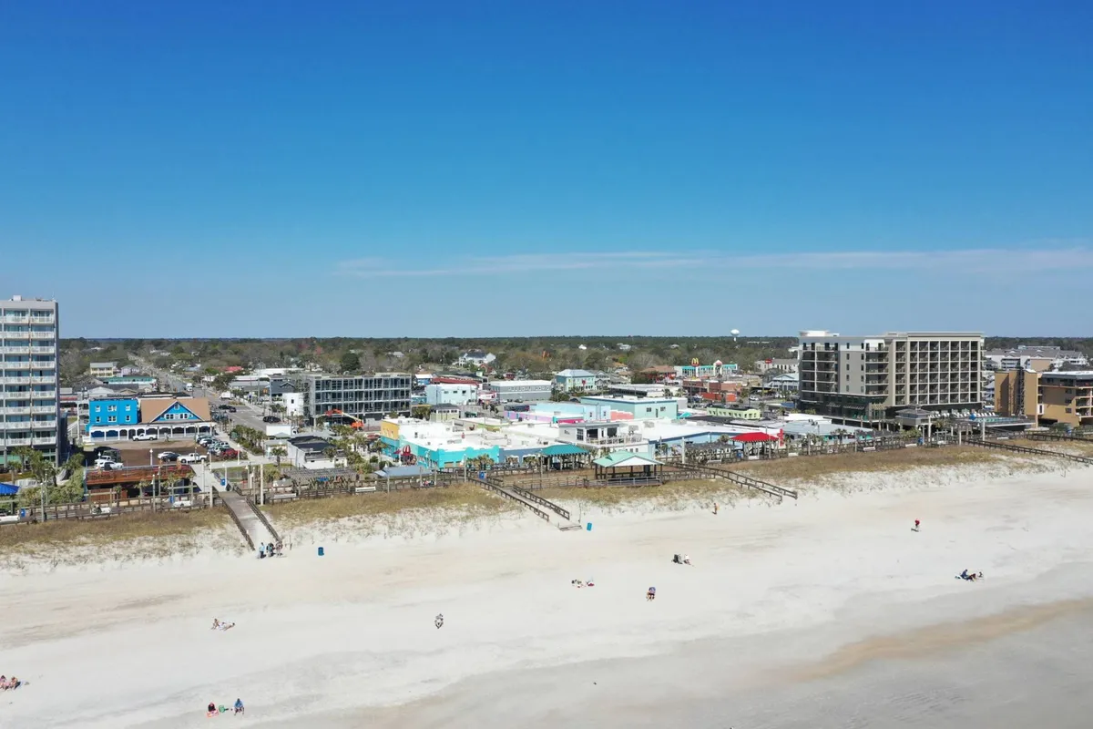 A vibrant exterior scene showcasing the Carolina Beach Boardwalk, colorful beachfront buildings, and the ocean. Families and visitors stroll, shop, and enjoy the lively boardwalk atmosphere, echoing the section's description of arcade games, donuts, and classic summer fun.