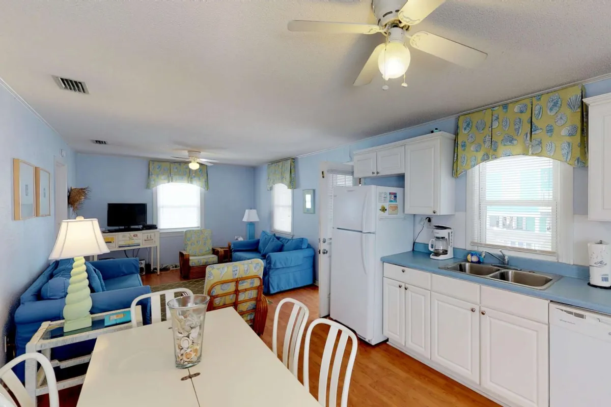 This interior shot focuses on a cozy living room with blue sofas, a dining table set for meals or games, and a kitchen counter nearby. Large windows allow sunlight to fill the space, reinforcing the airy, relaxed mood described in the section.