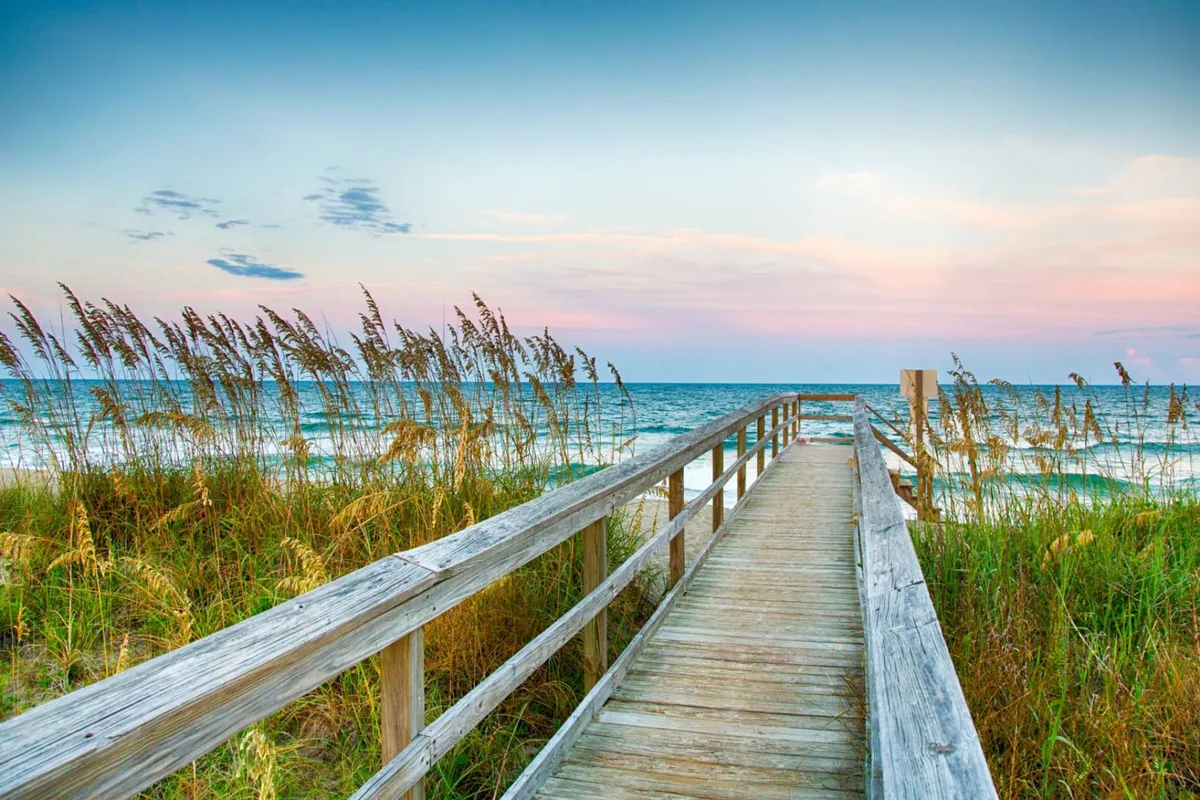 A wooden boardwalk stretches toward the ocean, bordered by seagrass and set against a colorful sunset sky. The scene captures the peaceful ambiance of a beach at dusk, with soft waves and the promise of a romantic stroll along the shore.