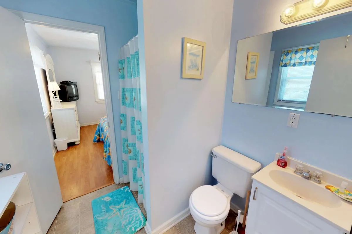 A bright Jack-and-Jill bathroom with a vanity, mirror, shower curtain, and a doorway leading to a bedroom. The atmosphere is serene and practical, ideal for shared use while maintaining privacy. The space is clearly designed for morning rituals and convenient routines.